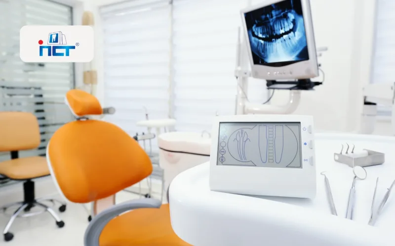 Dental clinic treatment room with orange dental chairs, white equipment, and a monitoring screen in the background.