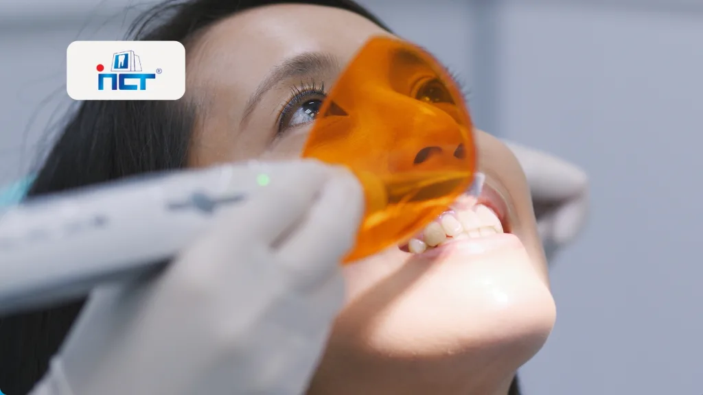 Dental patient with orange protective glasses during a dental exam, dentist's gloved hand near mouth; logo badge visible on the left