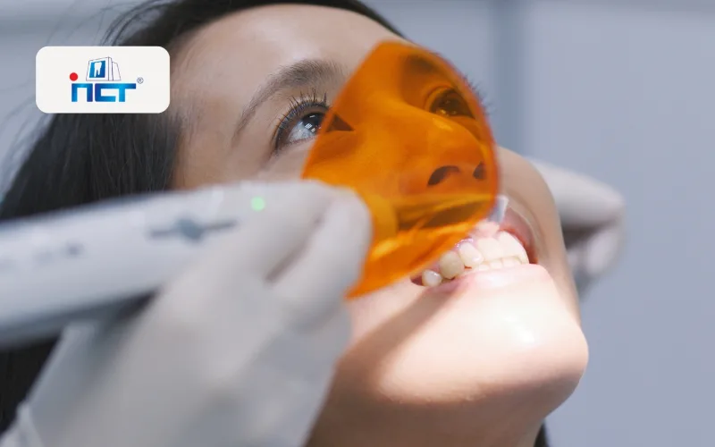 Dental patient with orange protective glasses during a dental exam, dentist's gloved hand near mouth; logo badge visible on the left