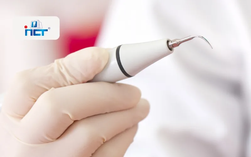 Gloved hand holding a dental scaler with a curved tip, ready for cleaning teeth (logo in background).